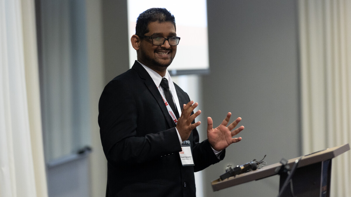 Revanth Raghupatruni, a brown man with black glasses, short black hair and a black moustache, wears a black suit while speaking at a podium.