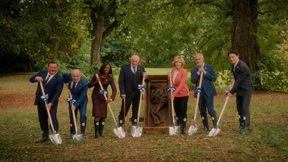 The seven main characters of the movie "Rumours" pose around a fossilized body in the woods.