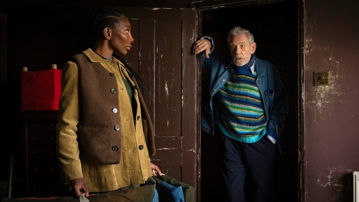Michaela Cole, a dark-skinned Black woman, stands on the left in a tan and brown tunic, next to a doorway where Ian McKellan, an older white man, stands in a green and blue striped sweater.
