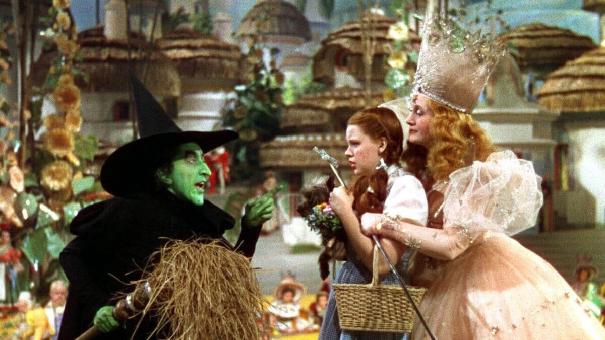 Dorothy, a white woman with long red hair, cowers in the arms of Glenda the Good Witch, a white woman in a pink dress and large tiara, in front of the Wicked Witch of the West, a green-skinned woman in a black jacket and pointy hat.