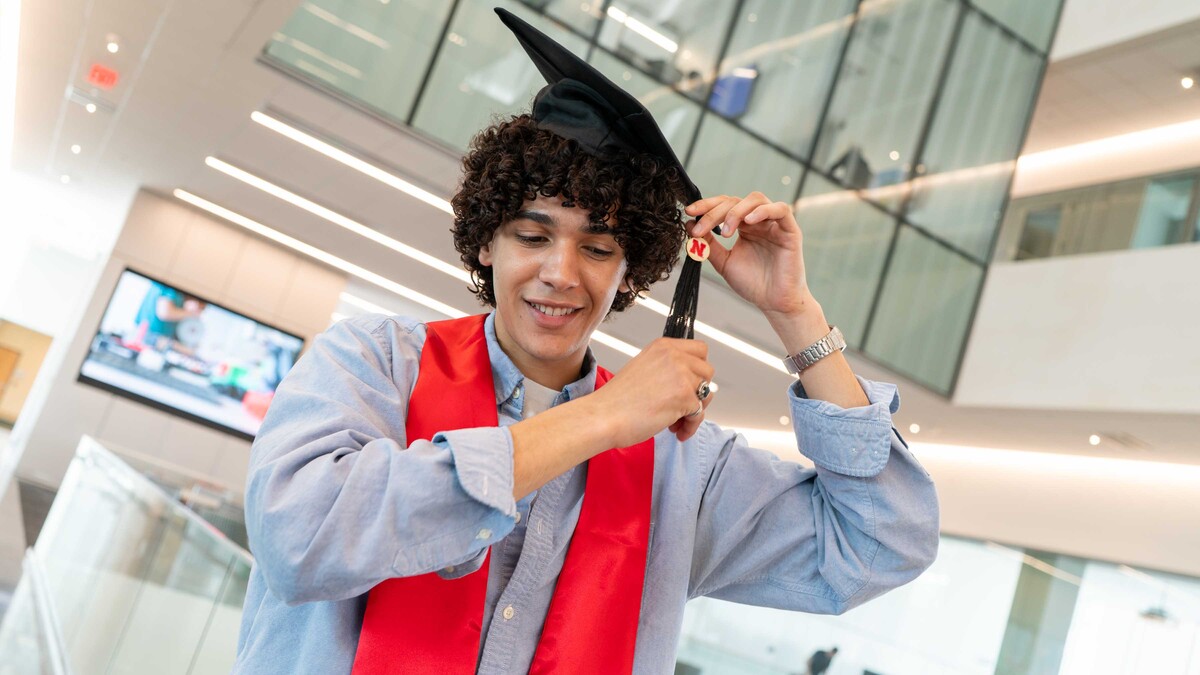 Adam Algahimi stands on a walkway in Kiewit Hall wearing blue button down shirt, a red graduation stole and black graduation cap. He adjusts the tassel on the cap, exposes the Nebraska 'N' button attached to it. 