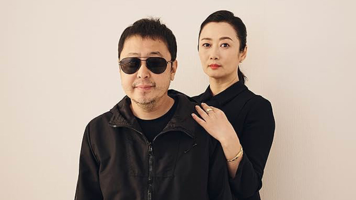 A man in a black jacket and sunglasses sits with a woman in a black sweater against a creme backdrop.