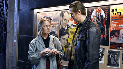 Image of a couple looking slightly confused as they stand in front of movie posters. The couple is featured in "Falling Leaves," a film playing Jan. 5-18 at UNL's Ross theater.