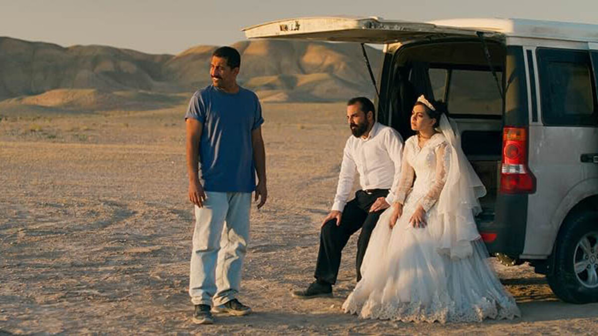 A brown man with dark hair, in a blue shirt and white pants stands with two people, a man and woman in wedding attire, resting on the back of a car.