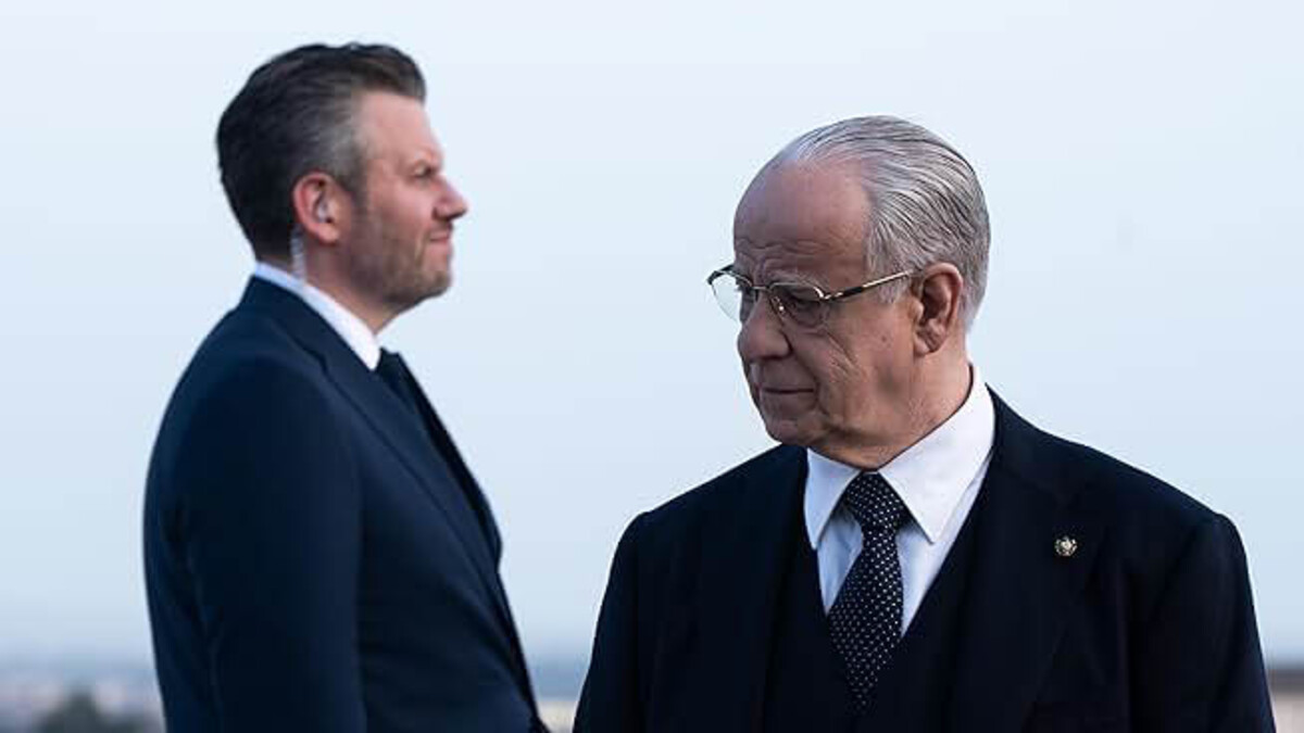 A brunette man in a black suit stands on the left, looking toward the right. An older, grey-haired man with glasses stands to the right, looking back at him.