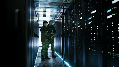 Two military men work in a hallway of server racks.