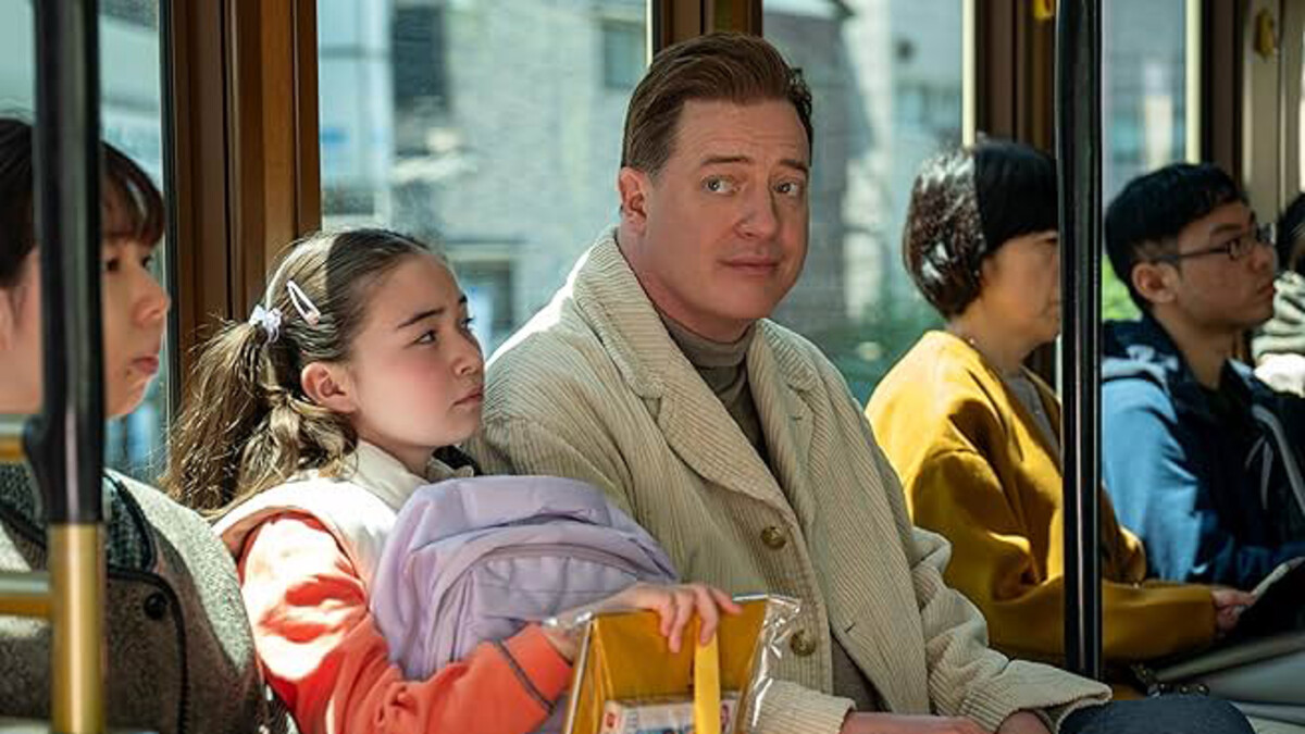 Brendan Fraser, a white man with short brown hair, sits in a creme coat with a young girl, who's long brown hair is in pigtails and arms are around her backpack.