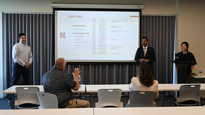 Multiple students in formal clothing stand around a tv, presenting their assignment for their supply chain management class.