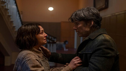 A brunette woman stands in front of a dirty brunette man in a hallway.