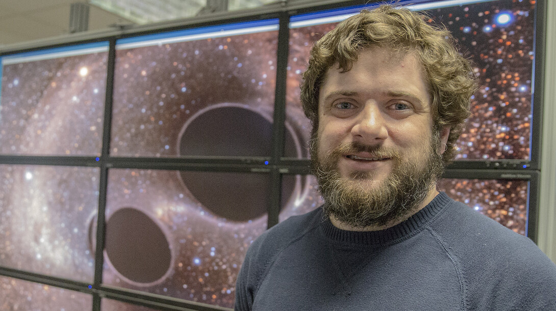 Brian Bockelman, research assistant professor of computer science and engineering, stands in front of a graphic that shows two black holes merging. Bockelman and colleagues assisted with the porting of code from LIGO's data facilities.