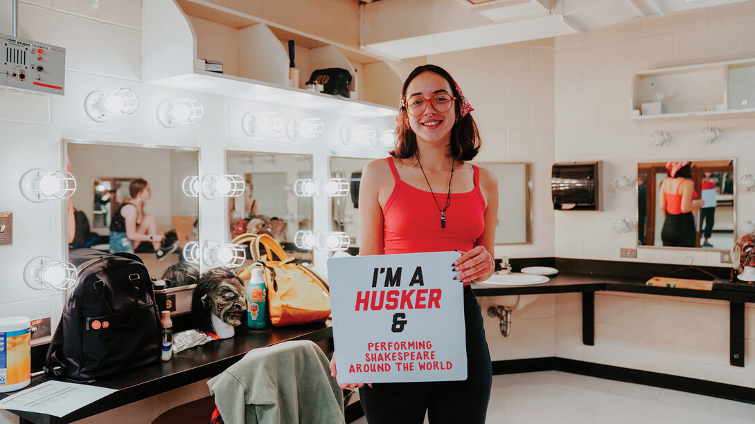 Standing in the dressing room of the Temple Building, Aurora holds up a sign that reads "I'm a Husker & Performing Shakespeare Around the World".