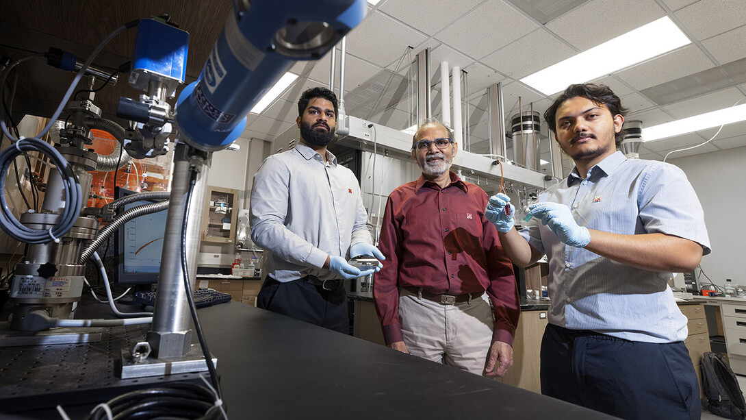 Abhijeet Prasad (left), a doctoral student in engineering; Ravi Saraf (center), Anderson Distinguished Professor of chemical and biomolecular engineering; and Aashish Subedi, a senior physics major, are photographed in Saraf’s Othmer Hall lab. Prasad is holding small electronic chips used to study the nanoparticle necklace network devices. Subedi is holding a suspension of nanoparticle necklaces (blue container) made from individual particles (red container).