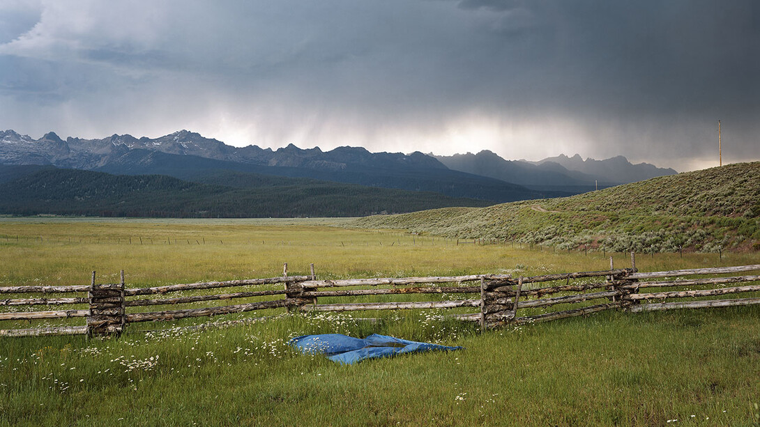 Green field with wooden fence in foreground and mountains in background, dark clouds dropping rain overhead