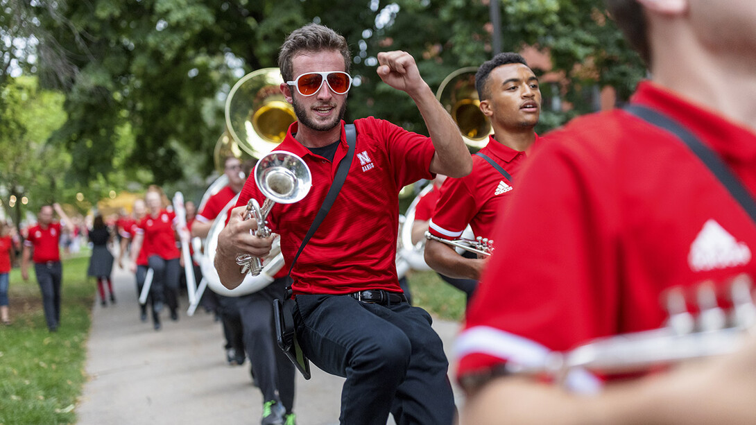 Cornhusker Marching Band members in red shirts and black pants march down the sidewalk near Kimball Hall