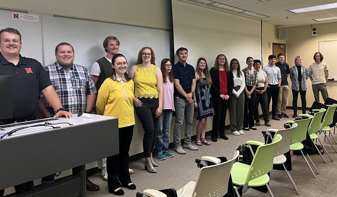 Fifteen Husker students stand at the front of a meeting room.