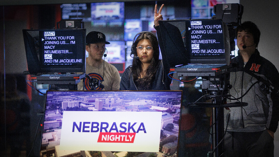 A young woman and two young men stand behind two teleprompters and a screen that reads "Nebraska Nightly."