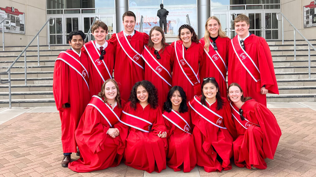 Twelve college students in red robes with red sashes pose on the plaza east of Memorial Stadium.