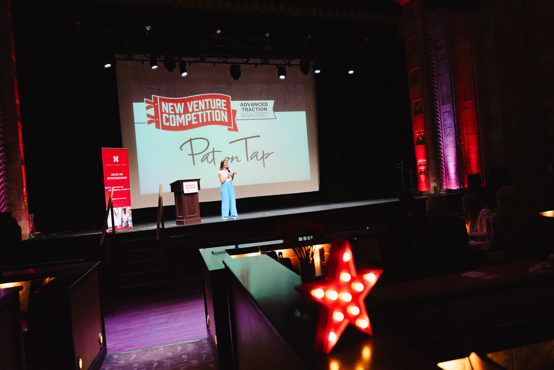 A young woman speaks on stage with a screen reading "Pat on Tap" in the background.