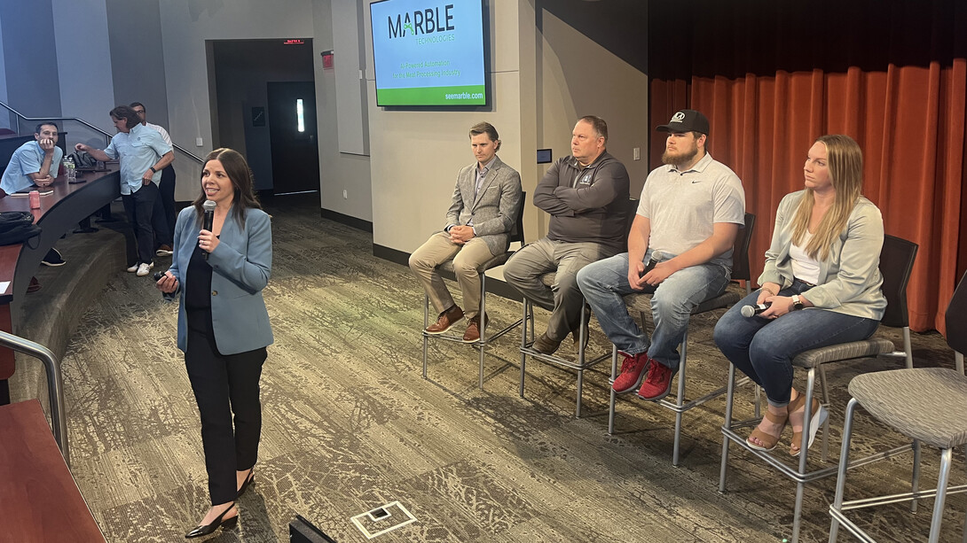 A woman with a microphone speaks while standing in front of three men and a woman seated in chairs.