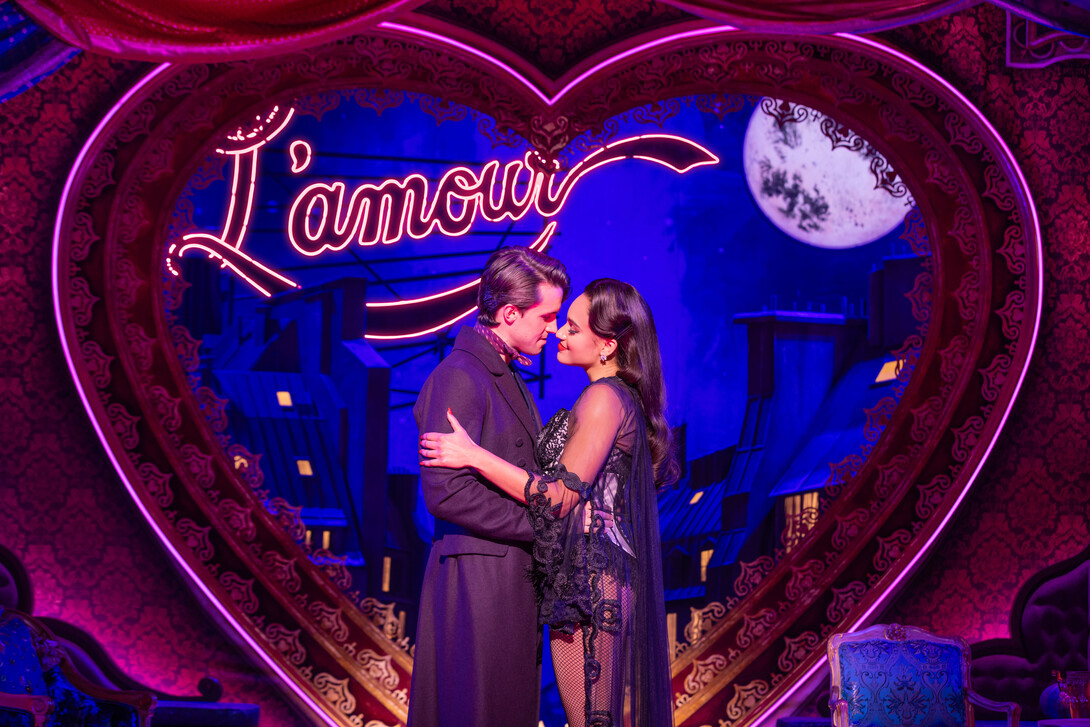 A young man, wearing a long coat and neckerchief, and young woman, wearing a black-and-white burlesque outfit, hold each other close in front of a nighttime cityscape outlined by a heart.