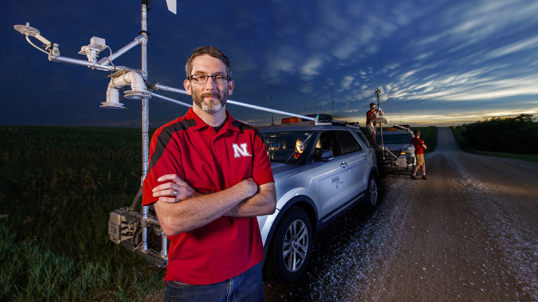 Adam Houston stands with his arms crossed in front of two storm-chasing vehicles.