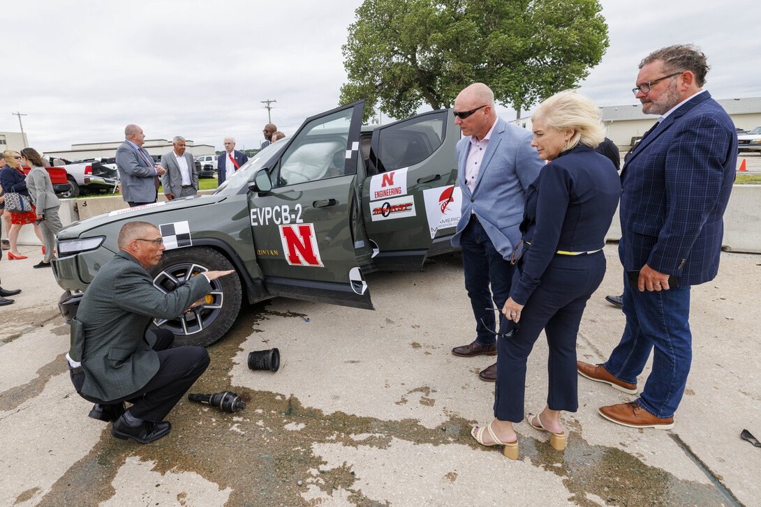 A man, crouching, speaks to two men and a woman, next to a crashed Rivian electric pickup.