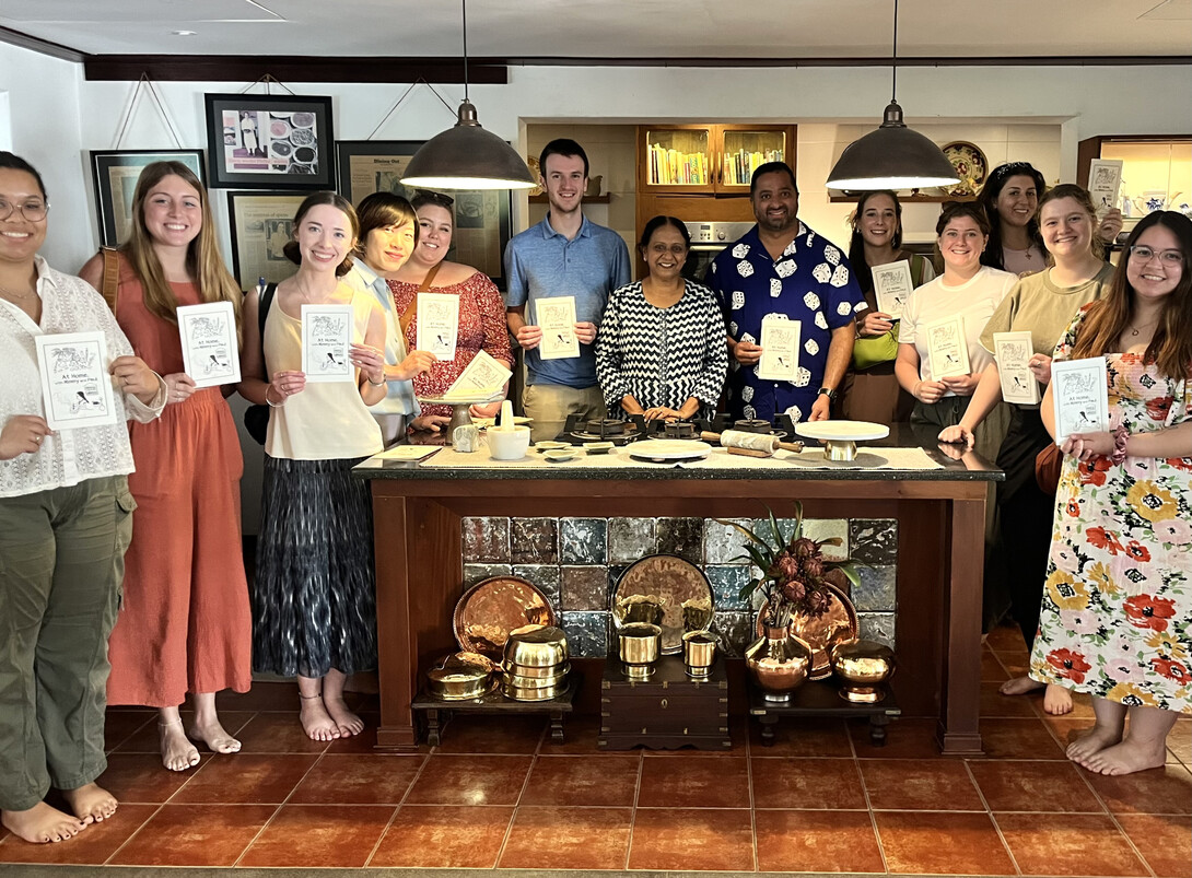 Fourteen people stand around a table in an Indian kitchen, many holding certificates.