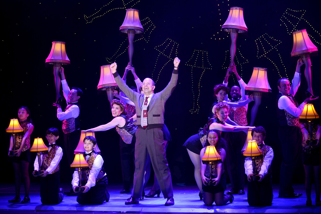 A middle-aged man in a sweater is surrounded by people wearing sparkly vests and bowties, carrying leg lamps, in this scene from "A Christmas Story: The Musical."