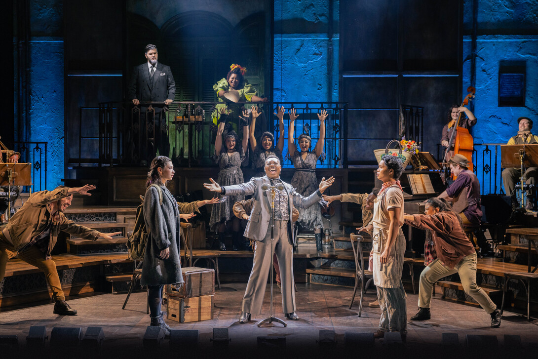 A man in a gray suit sings into a microphone, surrounded by a man and woman staring at each other, dancers, background singers and musicians in this scene from "Hadestown."