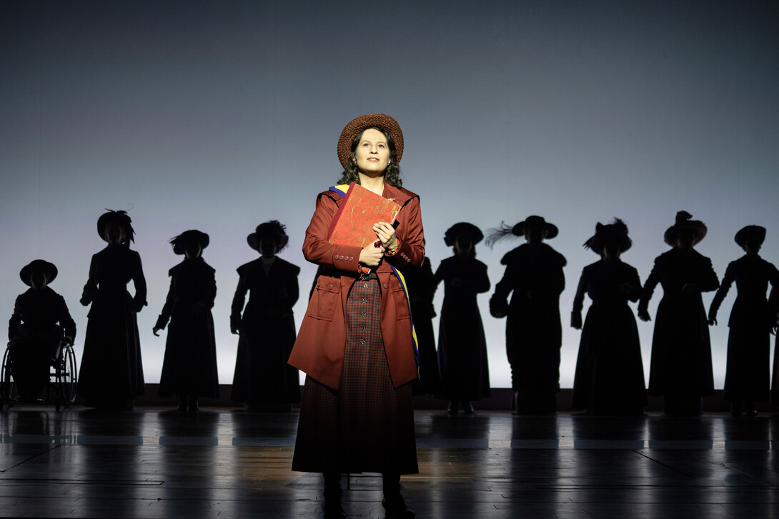 Shaina Taub (foreground) stars as women's rights activist Alice Paul in the musical "Suffs." She is seen in a maroon dress, overcoat and hat, holding a book, in front of a line of other suffragettes in silhouette.