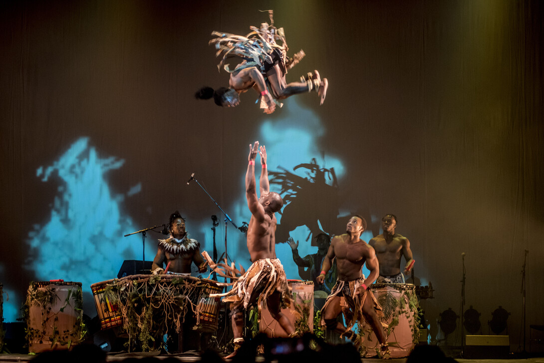 An acrobat prepares to catch another who is flipping in the air, near another acrobat and drummers.