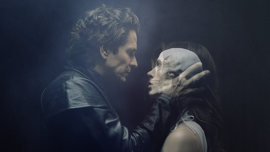 A man and woman stare at each other as he holds her head in his hands. A skull image shines through the woman's head.