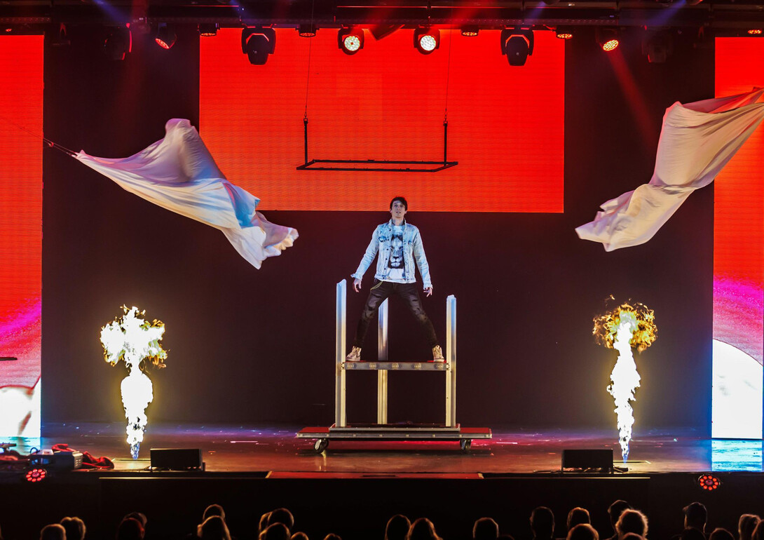 Magician REZA stands on a platform as two white sheets fly away from him and two flames erupt below him.