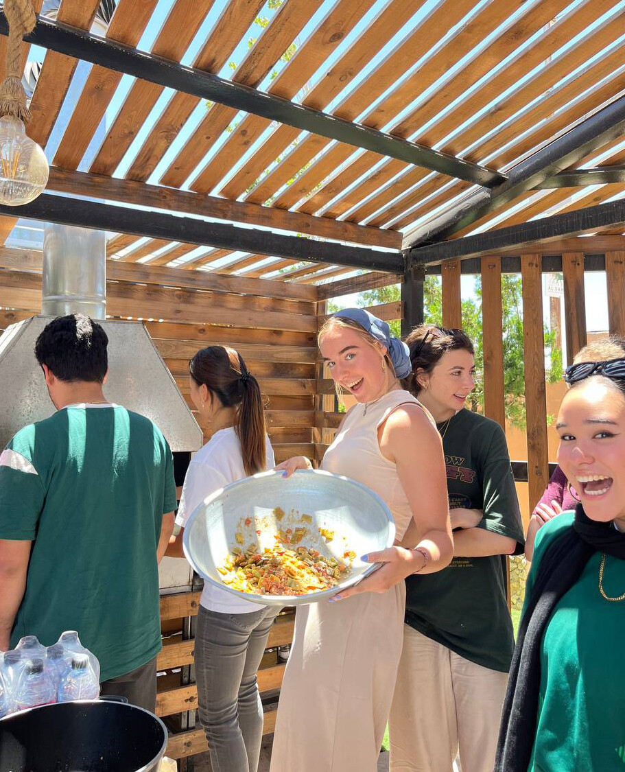 Kate Wandsnider smiles as she holds a large bowl of food in a wooden outdoor structure. Five other young people are near her.