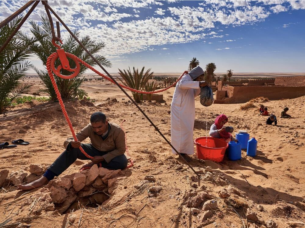 A man and two women use a manual pulley system to harvest water in the Algerian desert. Children sit in the sand nearby.