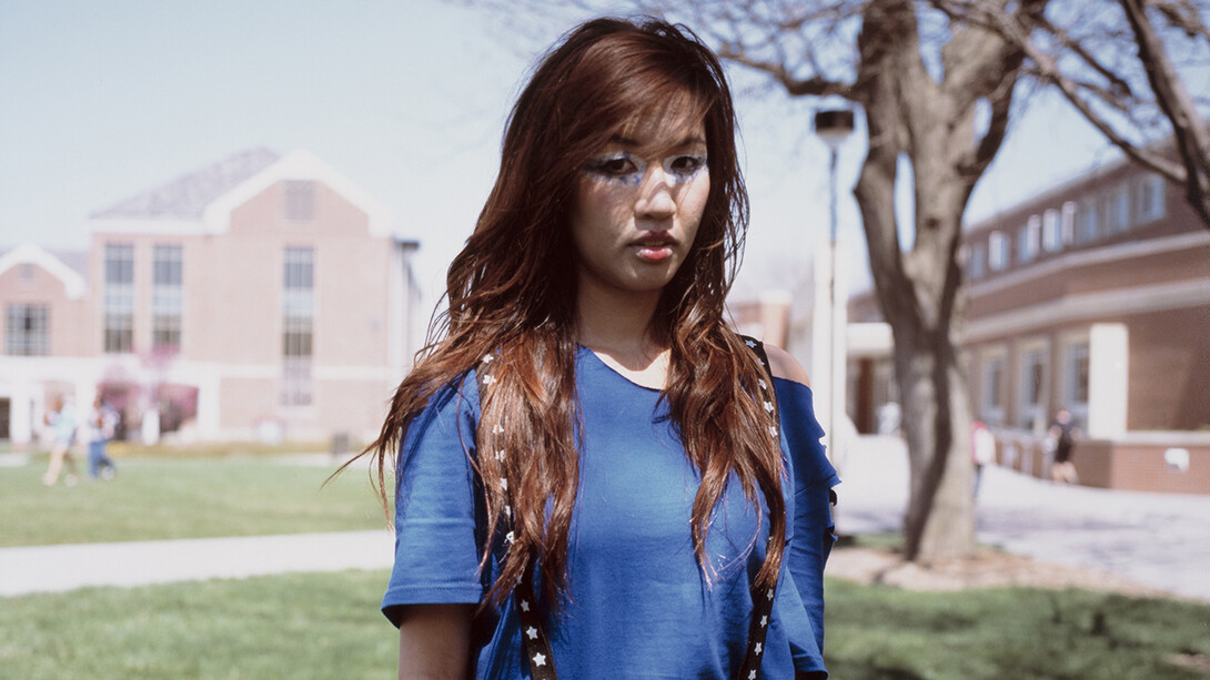 A posed photo of a young Vietnamese-American woman in front of the University of Nebraska–Lincoln's City Campus greenspace. She is wearing a blue shirt and black suspenders with white stars on them and has white-and-blue eye makeup.