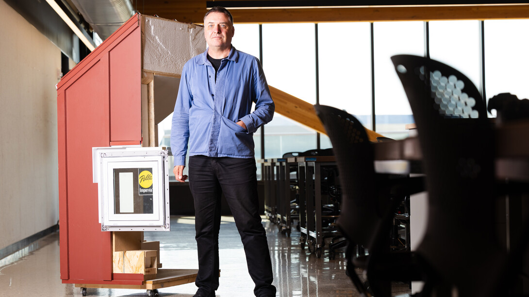 Jeffrey L. Day, professor of architecture at the University of Nebraska–Lincoln, stands in an architecture classroom. He is wearing a blue button-up shirt over a black shirt, black pants and black shoes. His left hand is in his shirt pocket.
