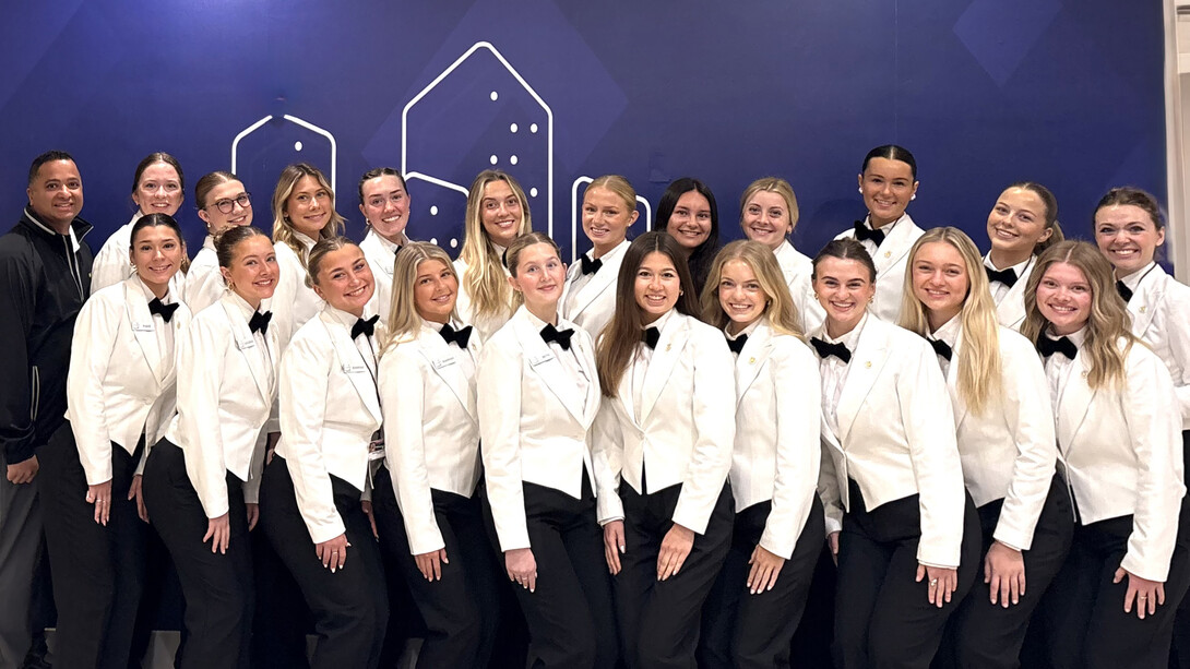 Fifteen young women wearing white jackets, white shirts, black bowties and black pants pose for a photo with instructor Ajai Ammachathram. They are Husker students providing hospitality services at the 2026 Masters Tournament.