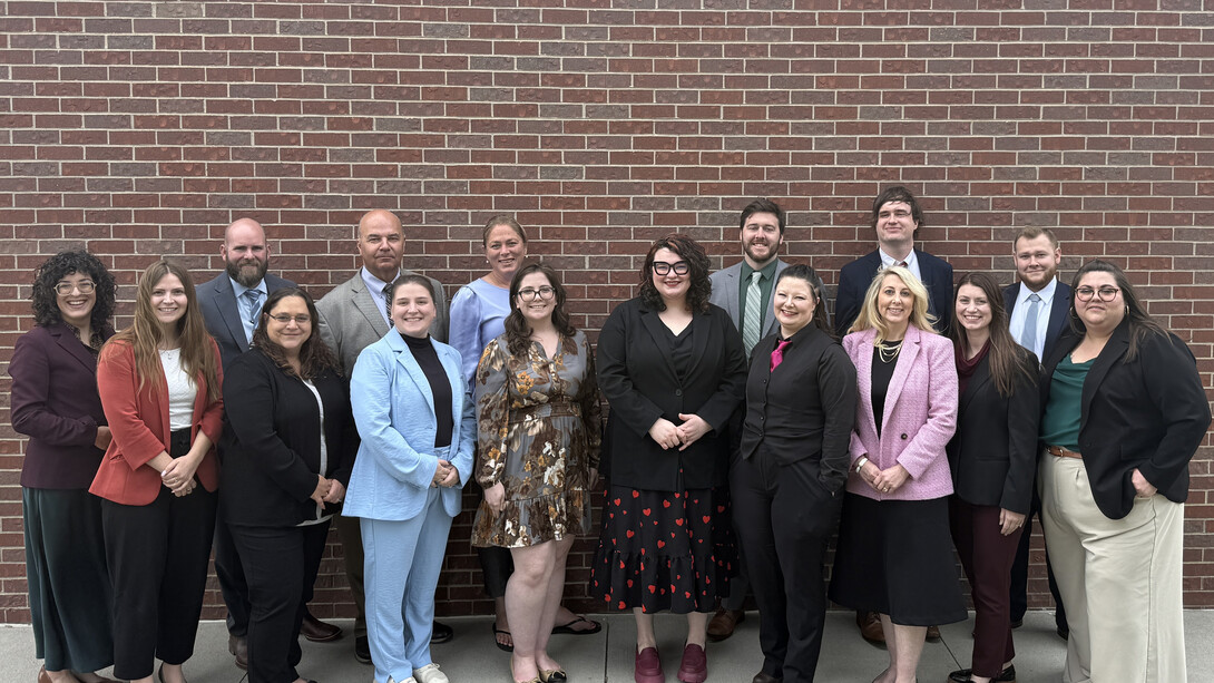 Sixteen attorneys in formal attire pose for a group photo in front of a brick wall.