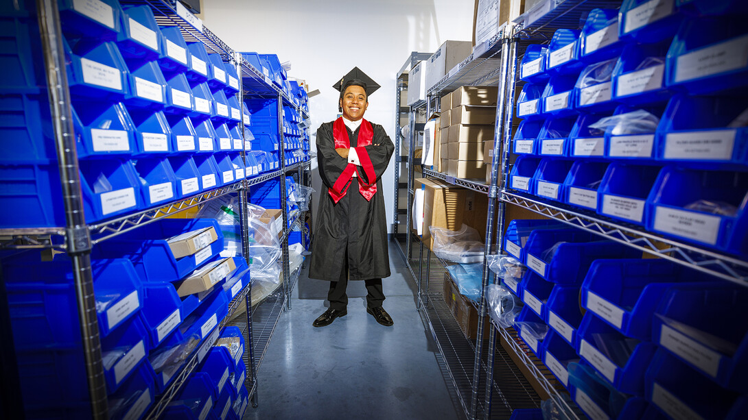 Brian Saravia Mejias in graduation regalia in a supply room.