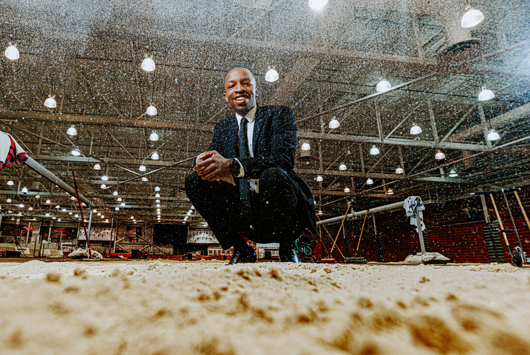 Nebraska's Passmore Mudundulu kneels at the edge of the jump pit in the Devaney Sports Center as sand rains down in front of the camera. Mudundulu is a junior marketing major from Lincoln.