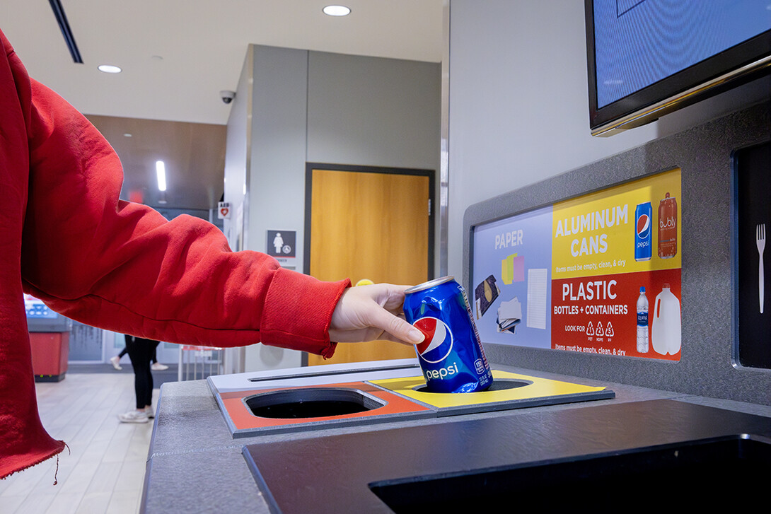 A hand places an aluminum can into an All in the Hall recycling station.