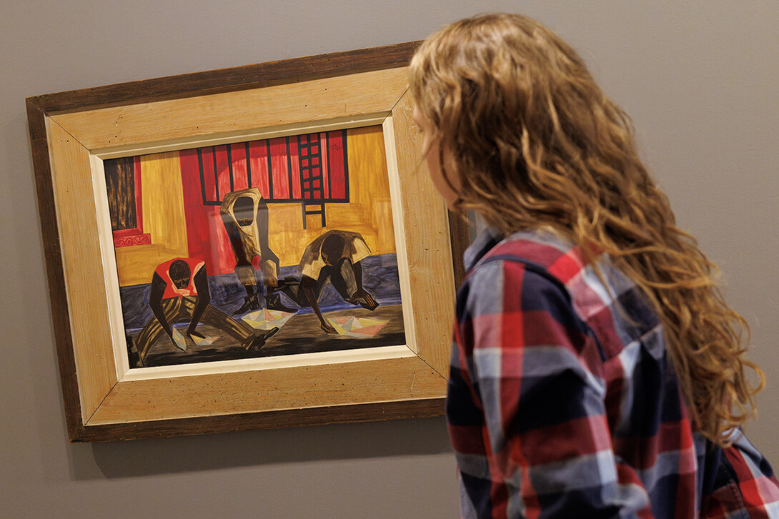 A person stands next to the framed painting "Paper Boats" by Jacob Lawrence