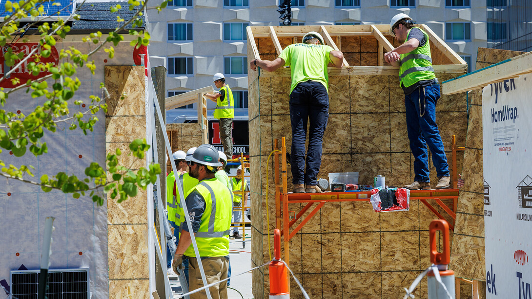 Two students in jeans, green reflective construction vests and white hard hats stand on an orange platform putting together two-by-fours while other students in the same attire work on other sheds around them.