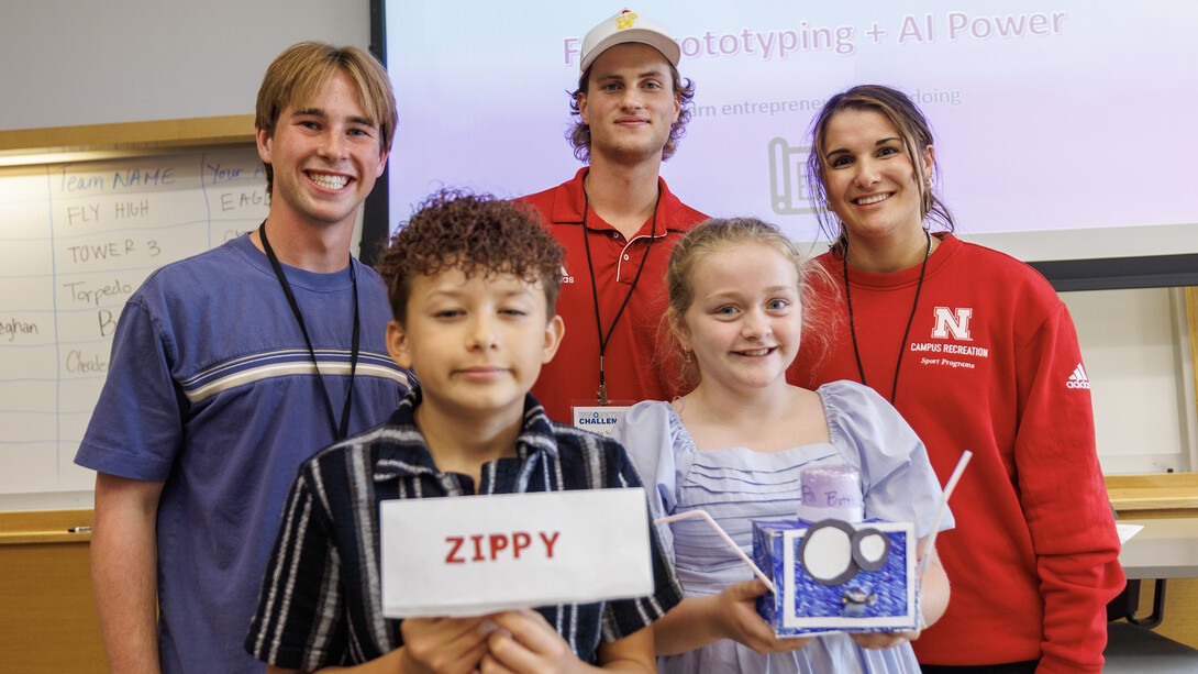 A blonde young man in a blue shirt, blonde young man in a white baseball hat and red shirt, and brunette woman in a red sweatshirt stand smiling with a brunette little boy with curly hair and a blonde little girl in a light blue dress.