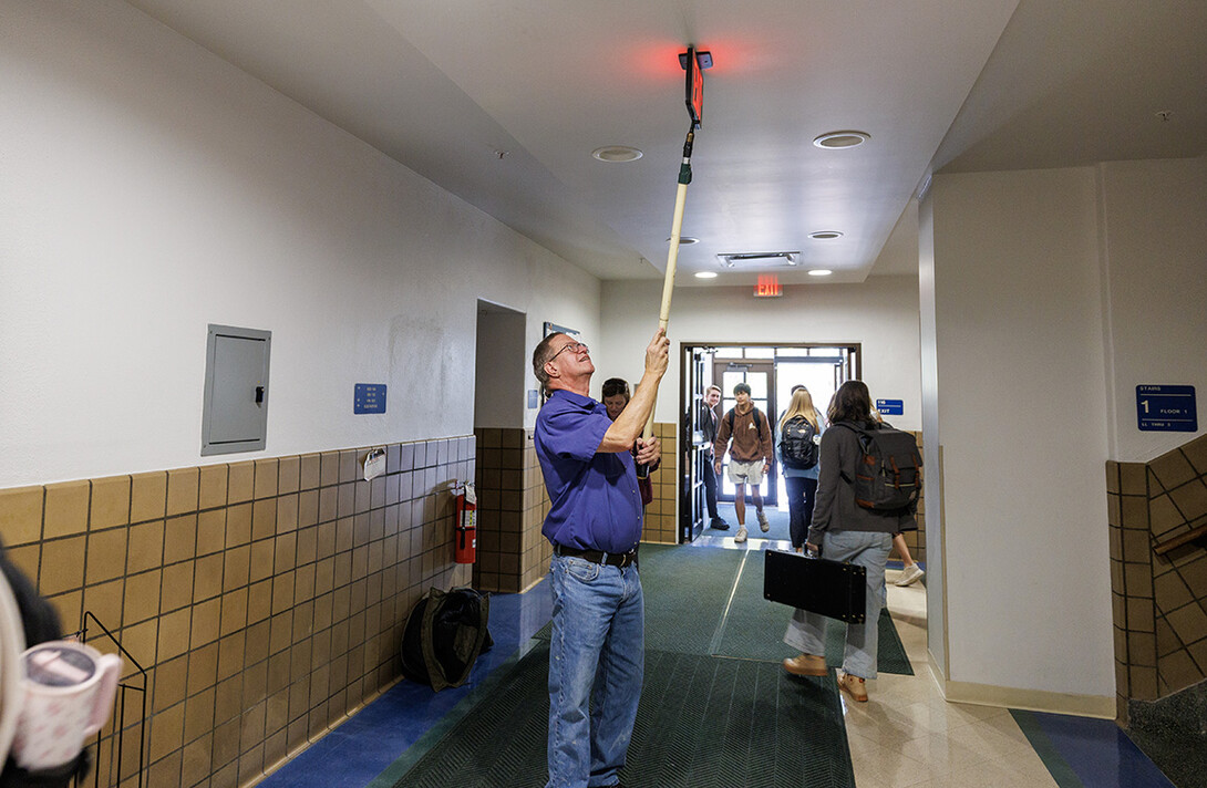 Duane Grosse, a unl Building Systems Technician, reaches for an exit sign in Burnett Hall to show how he uses his stick to test emergency lights.