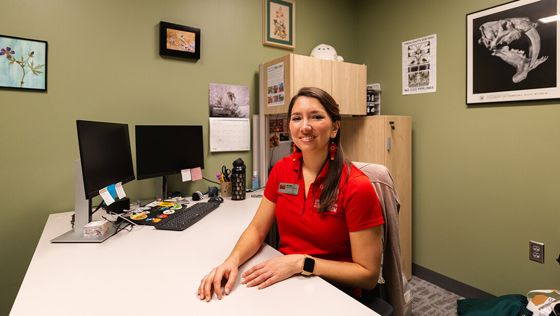 Ann Spilker, extended learning coordinator at the University of Nebraska State Museum, sits at her desk. Behind her is a large poster from the museum, and on the far left, a pressed flower made during one of Morrill Hall's paleobiology summer camp courses is framed over blue-painted paper.