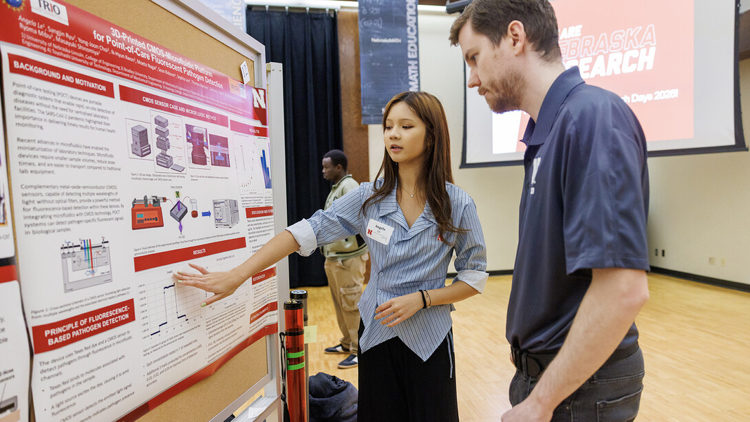 A student presents a research poster inside a large event space, speaking with attendees gathered nearby.