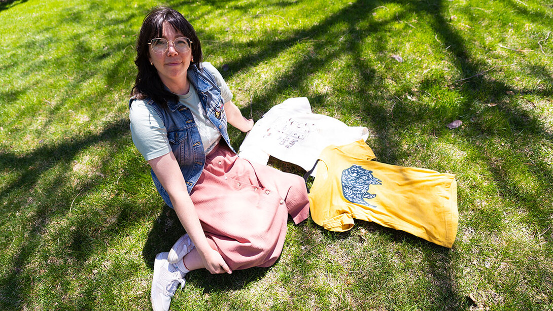 A woman sits next to two t-shirts on the grass.
