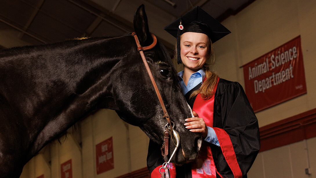 Izzi Velinsky, wearing a graduation cap and gown, stands next to a dark brown horse.